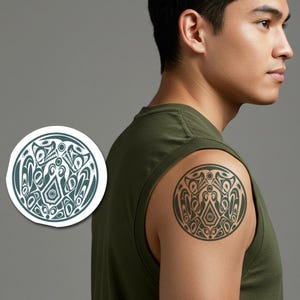 May include: A round, tribal-style tattoo design in dark gray ink on a person's shoulder. The design features symmetrical shapes and patterns within a circular border. A matching sticker is visible on the left.