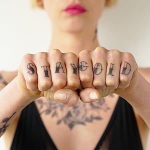 May include: A person's hand with the word "STAY GOLD" tattooed across the knuckles.