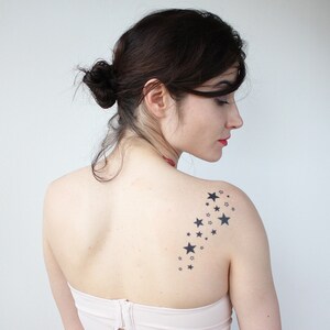 May include: A woman with dark hair pulled back in a bun has a temporary tattoo of a trail of black stars on her right shoulder.