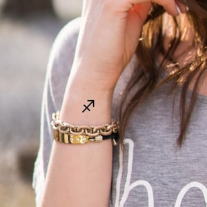 K&ouml;nnte beinhalten: Ein schwarzes tempor&auml;res Tattoo des Sternzeichen Sch&uuml;tze am Handgelenk einer Person.
