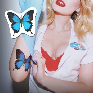 Puede incluir: Una mujer con un uniforme blanco con un sujetador rojo debajo y un tatuaje de mariposa azul en el brazo. El uniforme tiene una etiqueta con el nombre "blink-182".