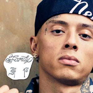 以下が含まれることがあります： 顔と首にタトゥーのある人物のクローズアップ。黒いキャップと金のイヤリングを着用しています。「Family 1st」という文字が入った白いステッカーが画像にあります。