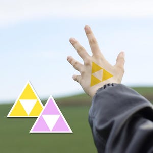May include: Two triangular stickers featuring a yellow and white design and a pink and white design, alongside a hand displaying a yellow and white triangular sticker on the back, all set against a blurred outdoor landscape background.
