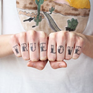 May include: A person's knuckles are tattooed with the words "TRUE LOVE" in a traditional style font.