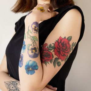 May include: A person's arm with temporary tattoos of flowers. The tattoos include red roses, purple pansies, and yellow flowers.