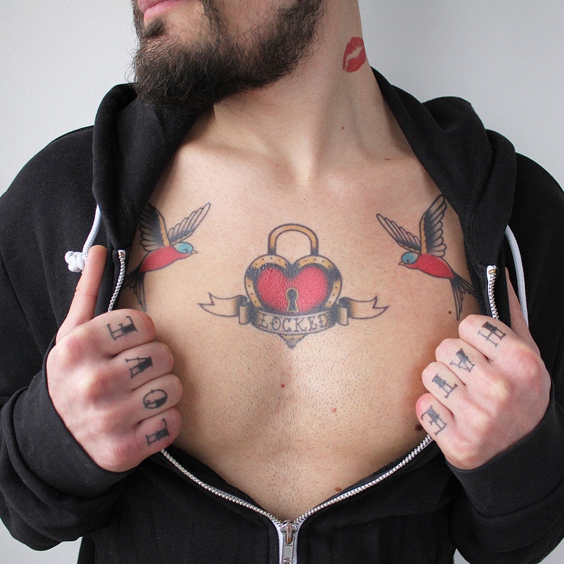 May include: A man with a beard has a tattoo on his chest. The tattoo is a red heart with a lock and key, with two red and blue birds on either side. The words "LOVE" and "HATE" are tattooed on his fingers.