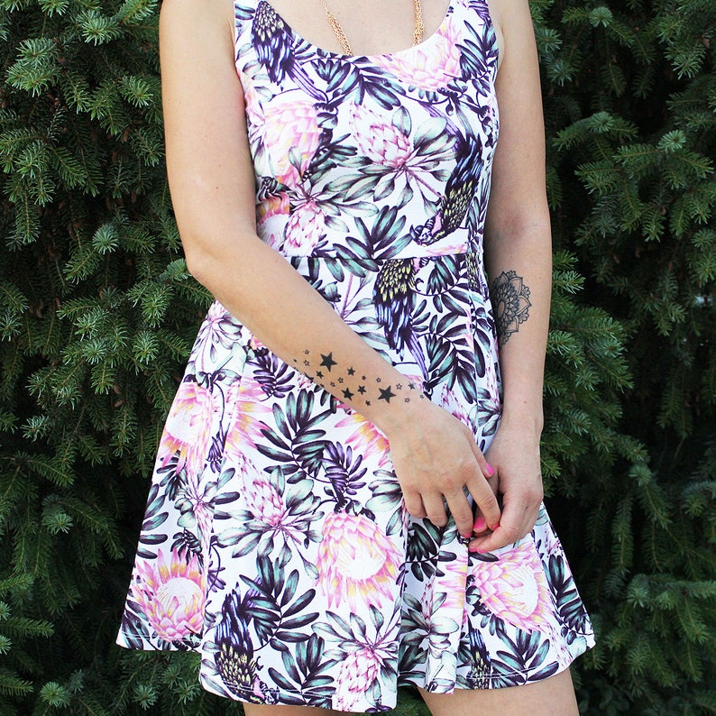 May include: A white dress with a floral print featuring pink, purple, and green flowers. The dress has a fitted bodice and a flared skirt. The model is wearing a gold necklace and has a star tattoo on her arm.