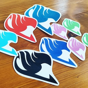 May include: A collection of colorful stickers featuring a stylized flame or wing design. The stickers are in various colors, including red, blue, black, pink, and green, and are arranged on a wooden surface.