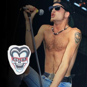 May include: A man wearing sunglasses and a backward baseball cap sings into a microphone. He has a tattoo of a skull with a heart-shaped outline on his arm. A patch with a skull wearing sunglasses is in the image.