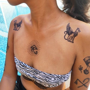 May include: A person with temporary tattoos on their chest and shoulders. The tattoos are black and include an eye with a sunburst, a winged cat, and a winged cat with a sunburst.