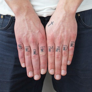 May include: A person's hands with the word "SELF MADE" tattooed on their fingers. A small cross tattoo is visible on the back of one hand.