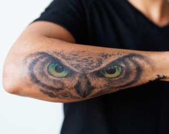 May include: A close-up of a person's arm with a black and white owl tattoo with green eyes.