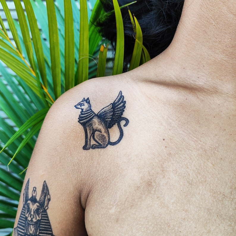 May include: A black and white tattoo of a winged cat on a person's shoulder.