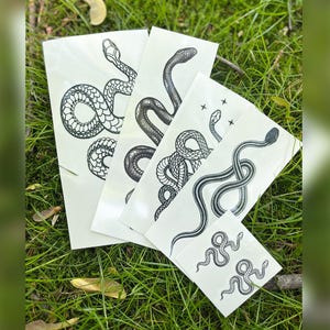 Puede incluir: Cuatro hojas de tatuajes temporales con diseños de serpientes negras. Los tatuajes están sobre un fondo blanco.