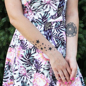 May include: A woman's arm with a temporary tattoo of black stars on her wrist. She is wearing a white dress with a floral print.