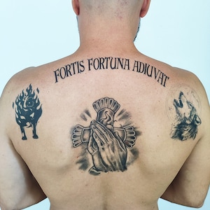 May include: Back tattoos featuring a cross with praying hands, a howling wolf, and a fiery creature. The phrase "FORTIS FORTUNA ADIUVAT" is arched across the upper back in black ink. The tattoos are detailed and shaded.