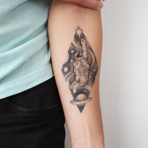 May include: A black and white temporary tattoo of an astronaut floating in space with a star, planets, and a galaxy swirling around them.