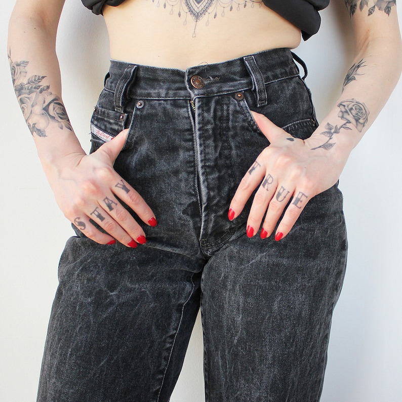 May include: A pair of black high-waisted jeans with a faded look. The jeans have a relaxed fit and are made of a soft, comfortable denim. The model is wearing the jeans with a black top and has tattoos on their arms that say "STAY TRUE".