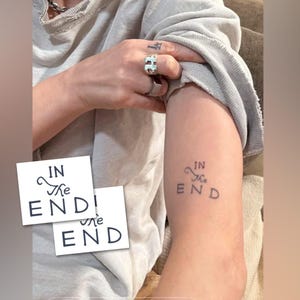 May include: A tattoo on an arm that reads "IN the END" in a stylized font. Two white cards with the same text are in the foreground. The person is wearing a gray sweatshirt and rings.