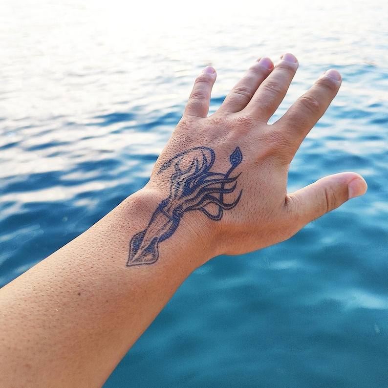 Puede incluir: Un brazo con un tatuaje temporal de un calamar en la mu&ntilde;eca. El tatuaje es negro y el calamar est&aacute; mirando hacia abajo.