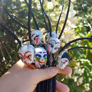 Puede incluir: Una colección de corbatas bolo con rostros de payaso de cerámica pintados a mano con diseños de maquillaje intrincados, engastes de metal plateado y detalles de cristal colgantes, unidos a cordones de cuero trenzado negro ajustables.