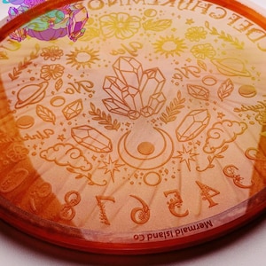 May include: An orange-tinted, round divination board with etched celestial and botanical designs. The board features crystals, planets, flowers, and the text "Mermaid Island Co." around the edge. A cartoon character with crystals is in the upper left corner.