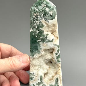 May include: A polished moss agate obelisk. The crystal features a mix of green, white, and tan colours. The obelisk is rectangular with a pointed top. The stone has a textured appearance with visible crystalline formations.
