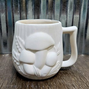 May include: A white ceramic mug with a textured surface and a large handle. The mug features a raised relief design of mushrooms and foliage. The mug is unpainted and ready for customization.