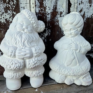 Könnte beinhalten: Zwei unbemalte Keramikfiguren: Weihnachtsmann und Frau Claus. Der Weihnachtsmann hat einen Bart und Hut, Frau Claus trägt eine Brille und strickt. Beide Figuren haben detaillierte Verzierungen und sind zum Bemalen bereit.