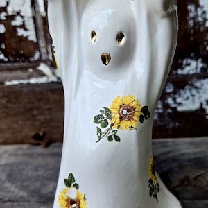 May include: White ceramic ghost figurine with raised arms and gold-colored eye accents. The ghost is decorated with yellow sunflowers and green leaves. The figurine is a decorative item.
