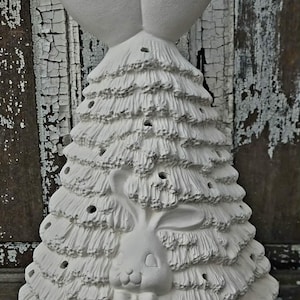 Peut inclure: Figurine de lapin de Pâques en céramique blanche. Le lapin est niché dans un motif texturé en forme d'arbre avec une base en forme de panier. Le lapin a de grandes oreilles et un nœud papillon. La figurine est non peinte et prête à être personnalisée.