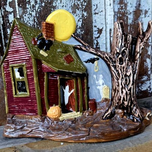 May include: A ceramic Halloween-themed display piece featuring a red house with a green roof, a yellow full moon, and a gnarled brown tree with a face. The scene includes a white ghost in the doorway, a pumpkin, a tombstone, and a black bat.