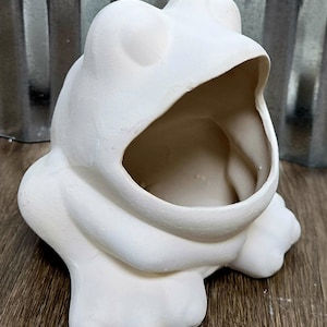 Könnte beinhalten: Ein weißer Keramik-Frosch-Pflanztopf mit weit geöffnetem Maul. Der Frosch hat große, runde Augen und sitzt auf einer Holzoberfläche. Der Pflanztopf ist unbemalt und bereit zur individuellen Gestaltung.