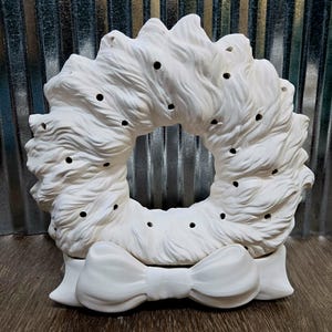 Könnte beinhalten: Ein weißer Keramik-Kranz mit einer Schleife am unteren Rand. Der Kranz hat ein strukturiertes, flauschiges Aussehen mit gleichmäßig verteilten Löchern. Die Schleife ist ebenfalls weiß und hat ein detailliertes, dreidimensionales Design. Der Kranz dient wahrscheinlich dekorativen Zwecken.