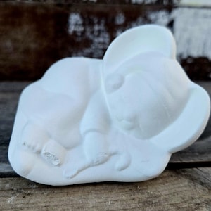 以下が含まれることがあります： 白いセラミック製の、帽子をかぶった眠っている赤ちゃんの置物。赤ちゃんは雲の形をした台座の上に丸まっています。置物は未塗装で、マットな仕上がりです。全体的なテーマはベビーデコレーションです。