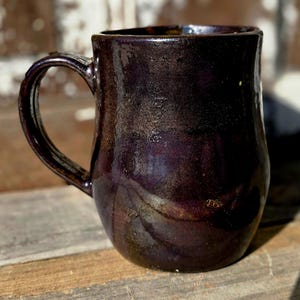 May include: A handcrafted ceramic mug with a dark, glossy finish. The mug has a deep purple hue with subtle brown and gold accents, and a curved handle. The mug is designed for hot beverages.