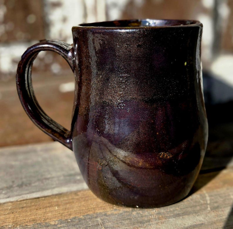 May include: A handcrafted ceramic mug with a dark, glossy finish. The mug has a deep purple hue with subtle brown and gold accents, and a curved handle. The mug is designed for hot beverages.