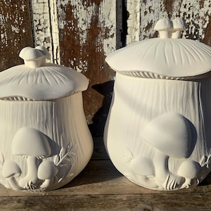 May include: Two unpainted ceramic mushroom-shaped jars with lids. The jars have a textured surface and are decorated with embossed mushroom and leaf designs. The lids are shaped like mushroom caps. The jars are set against a weathered wooden background.
