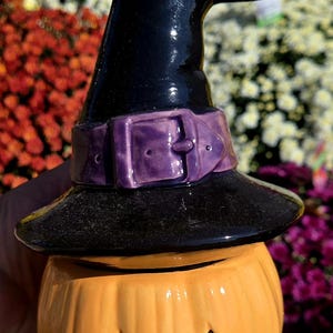 May include: A ceramic jack-o'-lantern jar with a glossy orange pumpkin base featuring a carved face. The lid is a black witch hat with a purple belt and buckle, topped with a small, light gray sculpted mouse figure peering from the top.