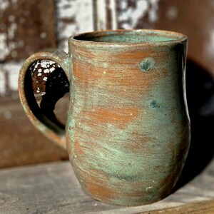 May include: Handmade ceramic mug with a rustic, textured finish. The mug features a blend of brown and teal-green hues, creating a unique, aged appearance. It has a large handle for easy gripping and is ideal for coffee or tea.
