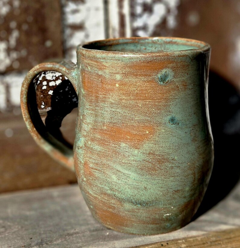 May include: Handmade ceramic mug with a rustic, textured finish. The mug features a blend of brown and teal-green hues, creating a unique, aged appearance. It has a large handle for easy gripping and is ideal for coffee or tea.