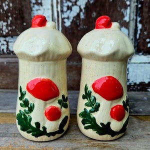Puede incluir: Dos saleros y pimenteros con forma de hongo. Los saleros y pimenteros son de color beige con tapas y detalles de hongos rojos. Hojas verdes y detalles adicionales de hongos rojos están pintados en los lados. Los saleros y pimenteros son de cerámica.