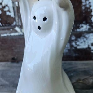 Könnte beinhalten: Eine weiße Keramik-Geisterfigur mit ausgestreckten Armen und schwarzen Punktaugen. Der Geist hat eine glatte, glänzende Oberfläche und einen abgerundeten Sockel. Diese Halloween-Dekoration ist ideal für eine gruselige Präsentation.