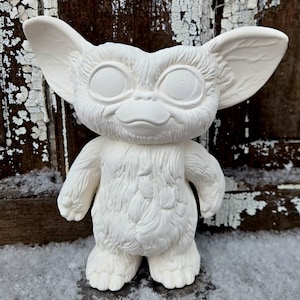 Op de afbeelding: Een witte keramische Gremlin-figuur met grote oren, een kleine neus en een harige borst. Het beeldje staat op een besneeuwd oppervlak, met een verweerde houten achtergrond.