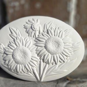 Puede incluir: Una placa ovalada de color blanco que presenta un relieve detallado de tres girasoles con centros texturizados y hojas puntiagudas. La superficie es mate y monocromática, resaltando el intrincado diseño floral mediante profundidad y sombra.
