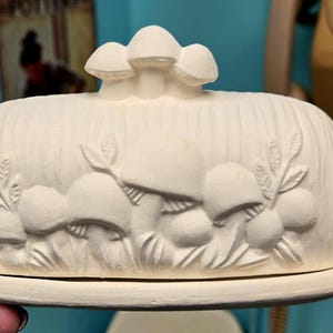 Puede incluir: Mantequera de cerámica blanquecina con tapa, con un diseño en relieve de setas y follaje. La mantequera tiene una base rectangular y una tapa abovedada, coronada con tres esculturas de setas. Medidas en centímetros.