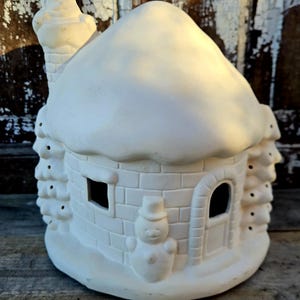 May include: A ceramic, off-white, winter-themed house with a snow-covered roof, chimney, and a snowman. The house has a door, a window, and decorative trees on the sides. The house is made of brick-like blocks.