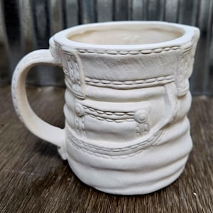 Könnte beinhalten: Eine weiße Keramiktasse mit Henkel, die ein strukturiertes Design aufweist, das einer Jeans ähnelt. Die Tasse hat detaillierte Nähte und Knopfakzente, wodurch ein einzigartiges, handgefertigtes Aussehen entsteht. Die Tasse ist unbemalt und bereit zur Individualisierung.