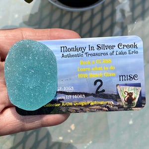 May include: A piece of sea glass in a light blue hue, held next to a business card. The card reads "Monkey In Silver Creek" and "Authentic Treasures of Lake Erie". The card also has text that says "Book a CLASS" and "Learn what to do With Beach Glass".