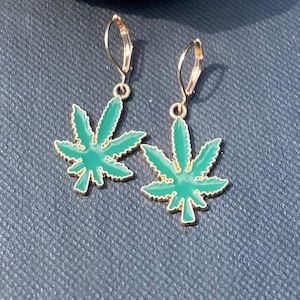 May include: A pair of gold-toned leverback earrings featuring dangling charms shaped like seven-pointed cannabis leaves. The leaves are filled with a vibrant green enamel finish, and the metal edges are polished to a bright gold-toned shine.
